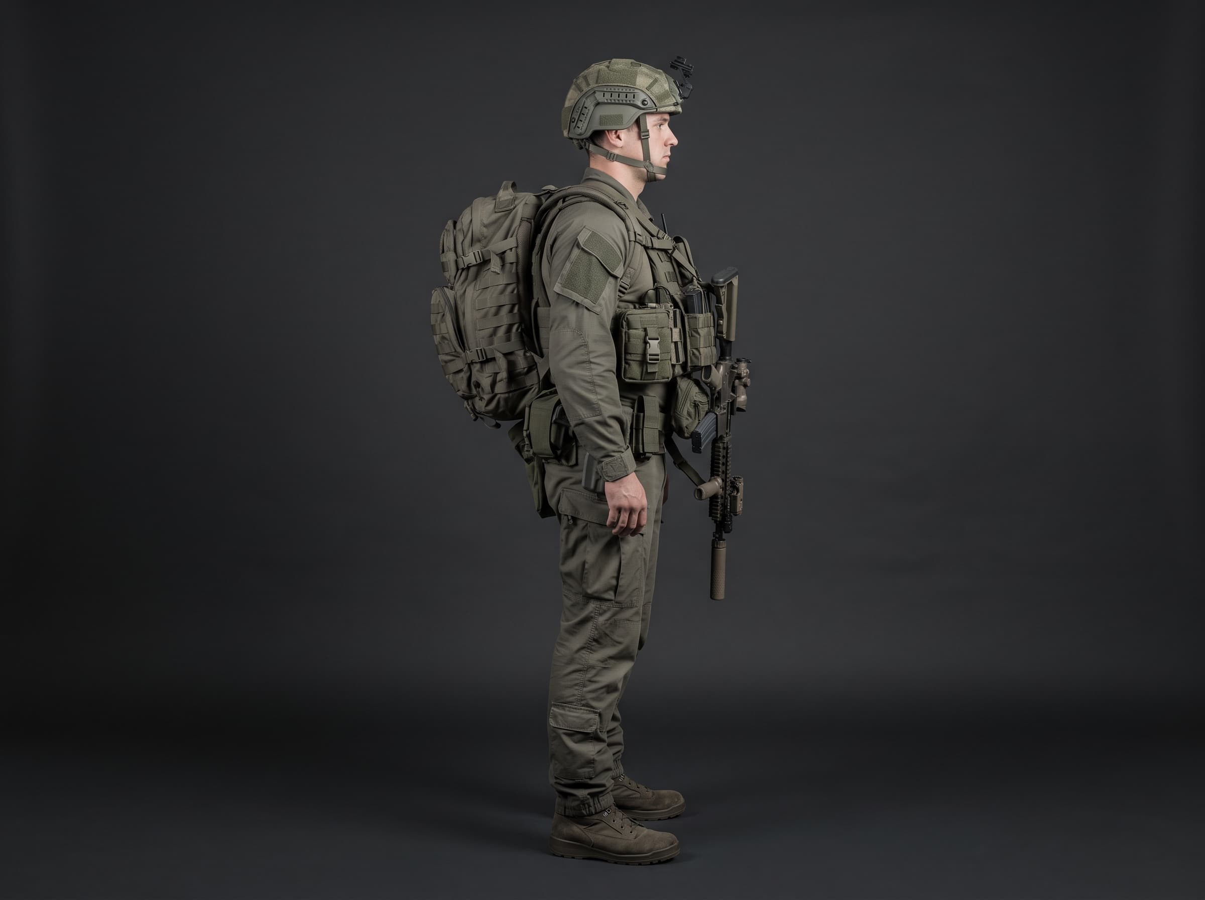 Side profile of a soldier standing in full kit — helmet, backpack, plate carrier, rifle and boots — against a dark studio background