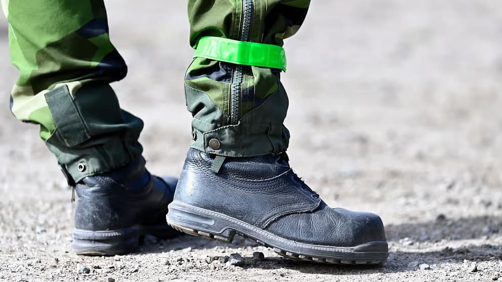 Close-up of a conscript's legs in Swedish camouflage trousers and black leather boots on gravel, a green reflective band around one ankle